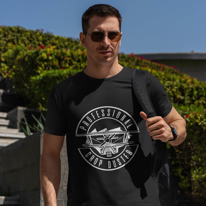 Man wearing sunglasses and a black "Professional Crop Duster" t-shirt, holding a backpack strap, standing outdoors near greenery and concrete steps.