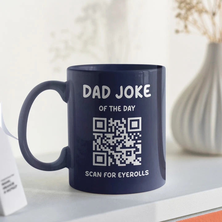 A navy blue mug with the text "Dad Joke of the Day" above a QR code and the words "Scan for eyerolls" printed below. A white vase with dried flowers is in the background.