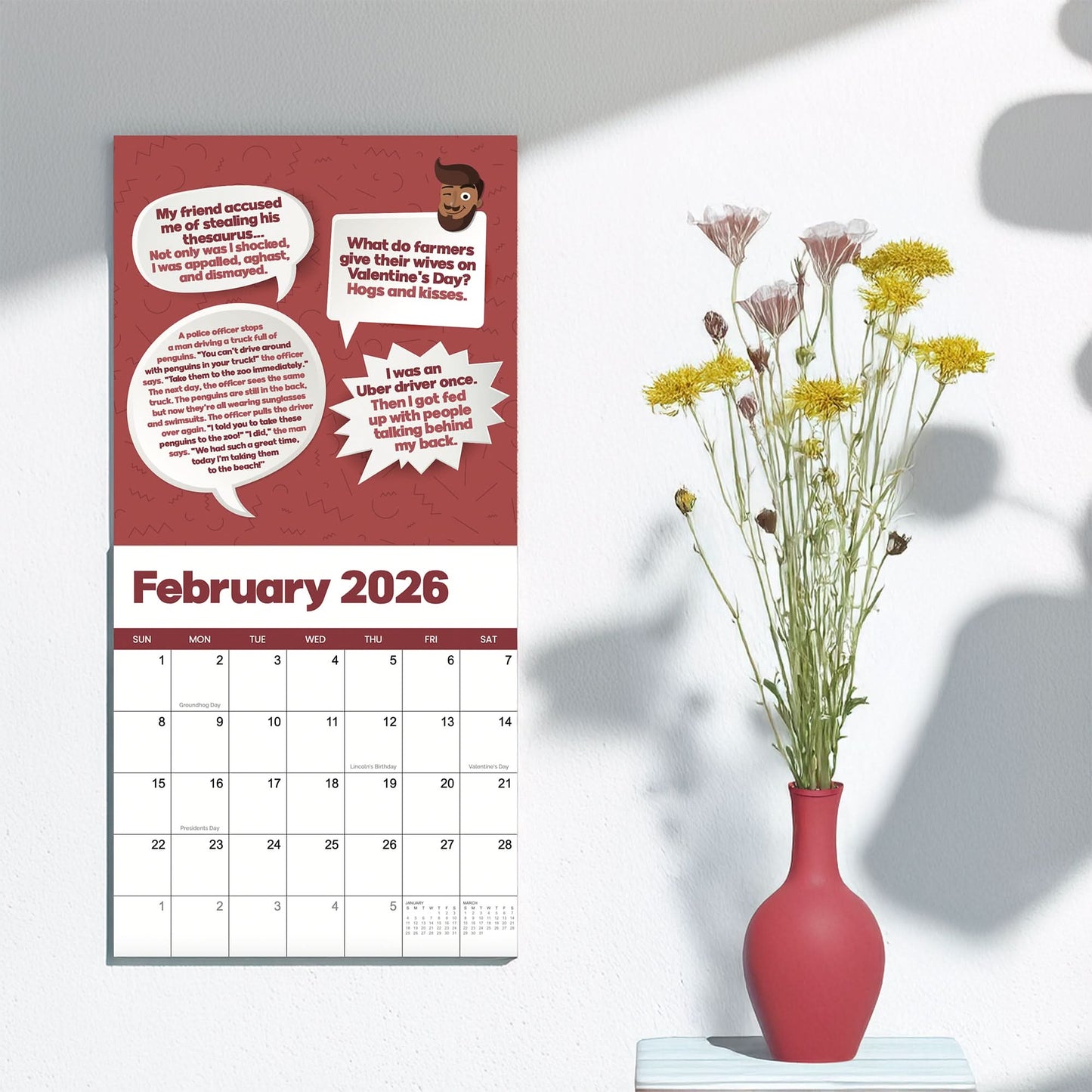 A 2026 Dad Joke Wall Calendar by That Is So Dad hangs open to February next to a white table with a red vase filled with yellow and white flowers.