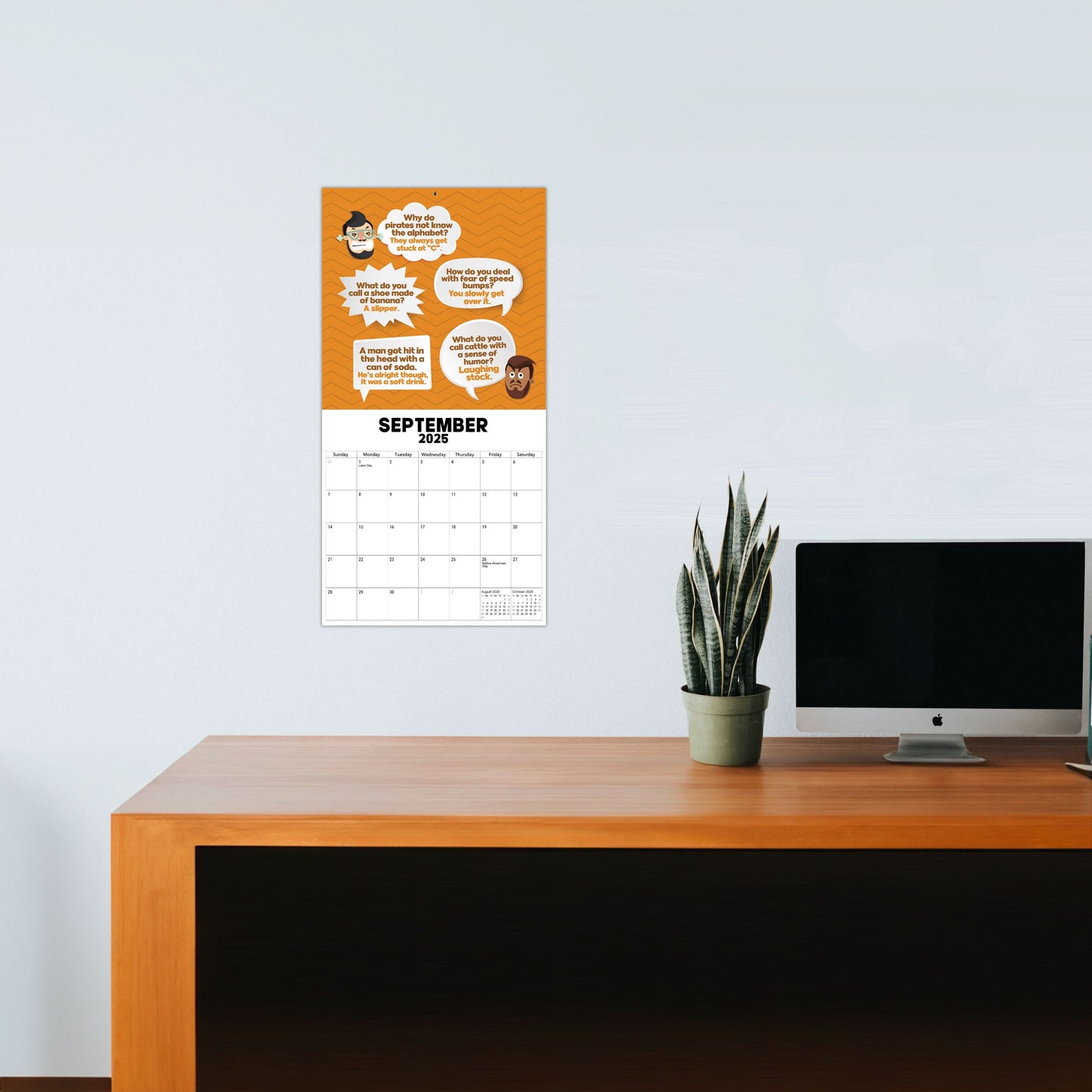 A wooden desk with a potted plant and a monitor, and on a white wall hangs the "2025 Dad Joke Wall Calendar" by That Is So Dad, featuring speech bubbles with quotes.