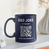 A navy blue mug with the text "Dad Joke of the Day" above a QR code and the words "Scan for eyerolls" printed below. A white vase with dried flowers is in the background.
