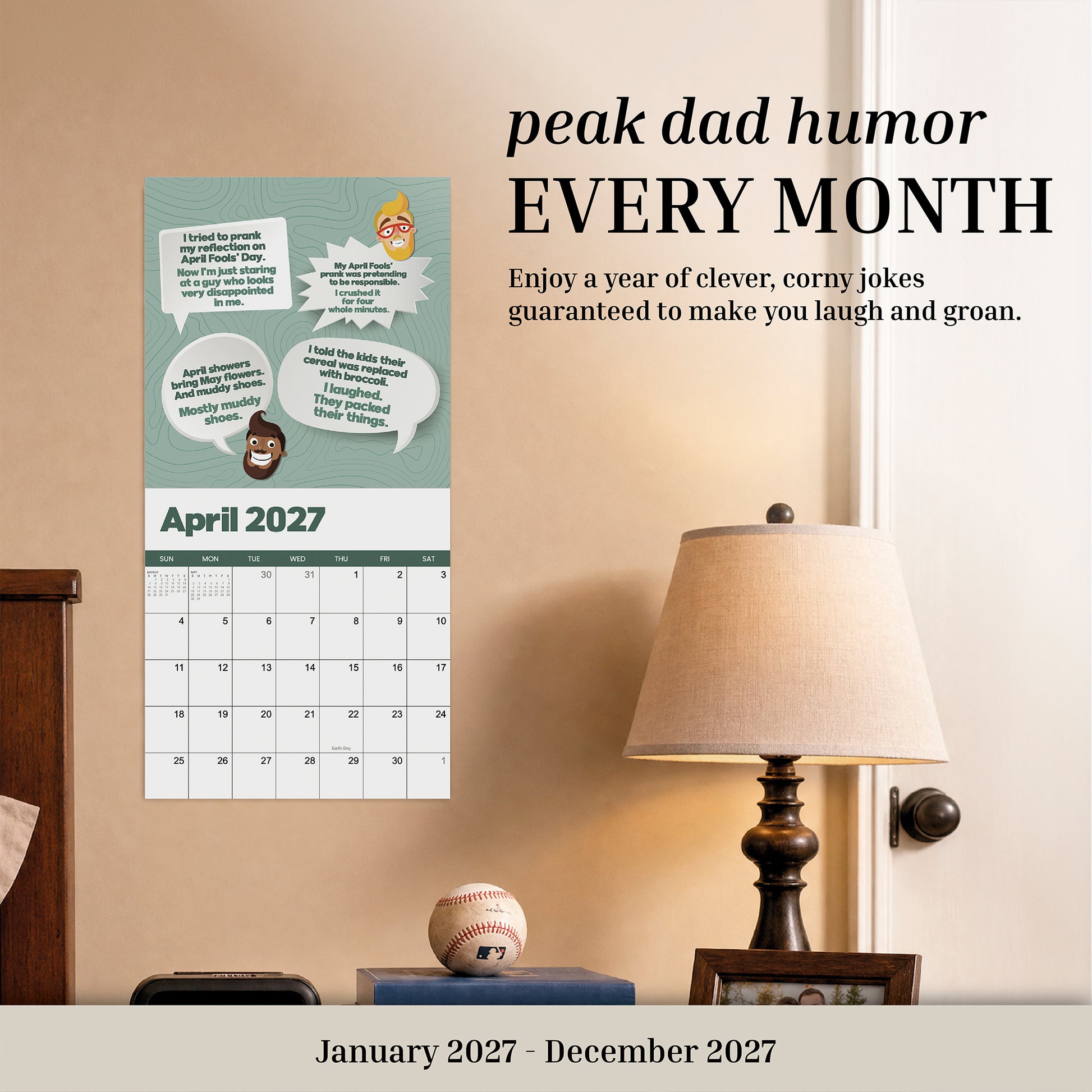 A 2027 Dad Joke Wall Calendar from That Is So Dad hangs above a nightstand with a lamp, baseball, and clock. This humorous calendar features dad joke cartoons for the month of April.