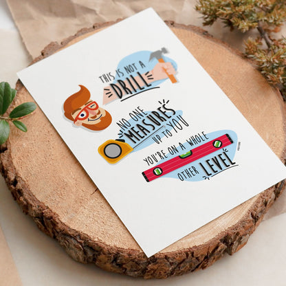 The "This Is Not A Drill" Funny Father's Day Card by That Is So Dad sits on a wooden slab, showing a man, drill, tape measure, and level—ideal for Father’s Day with its clever message about measuring up and leveling up.