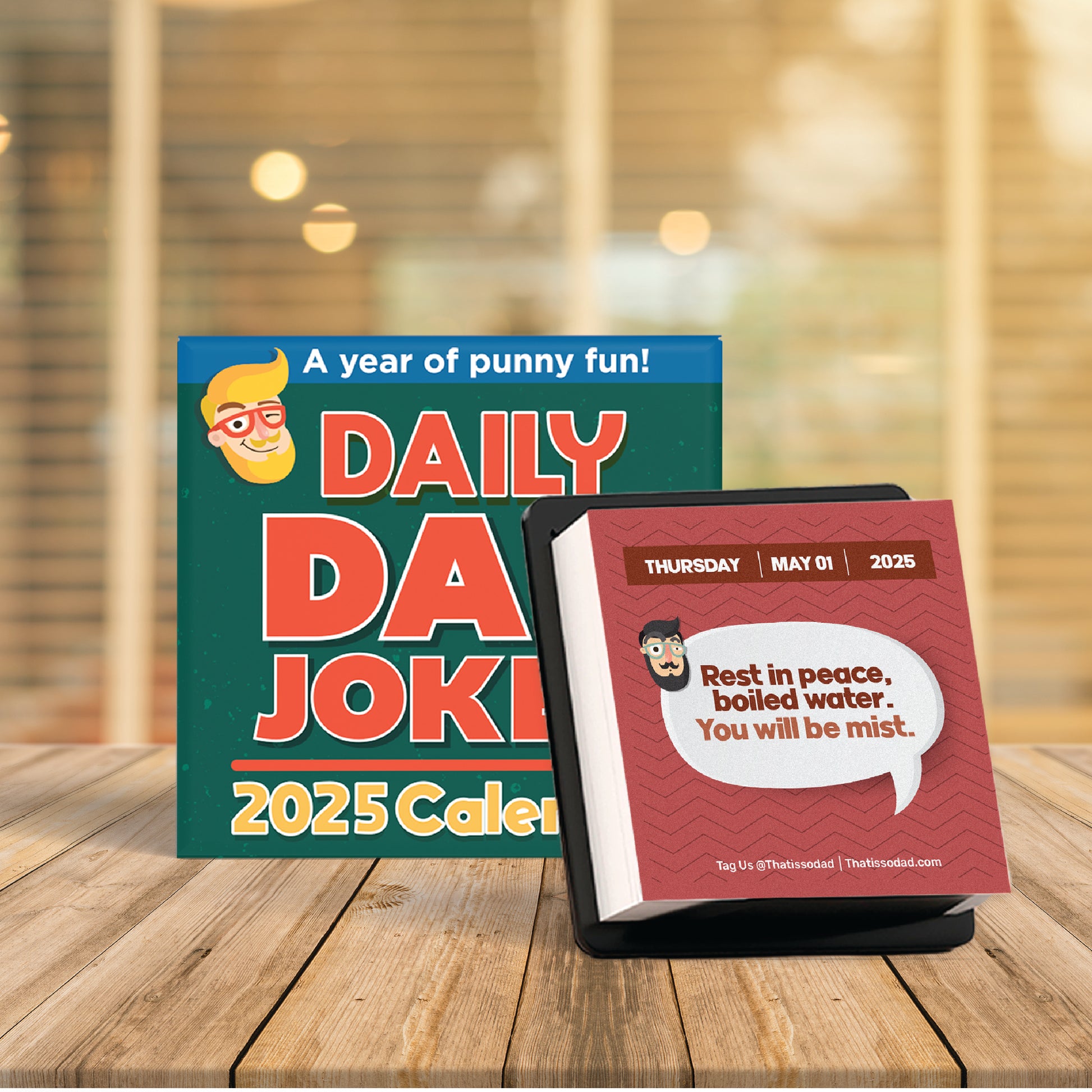 A 2025 Dad Joke Gift Set by That Is So Dad, featuring a desktop calendar complete with an easel backer for easy display, sits on a wooden table. The page for May 1 showcases the hilarious dad joke: "Rest in peace, boiled water. You will be mist." All of this is set against a beautifully blurred background.