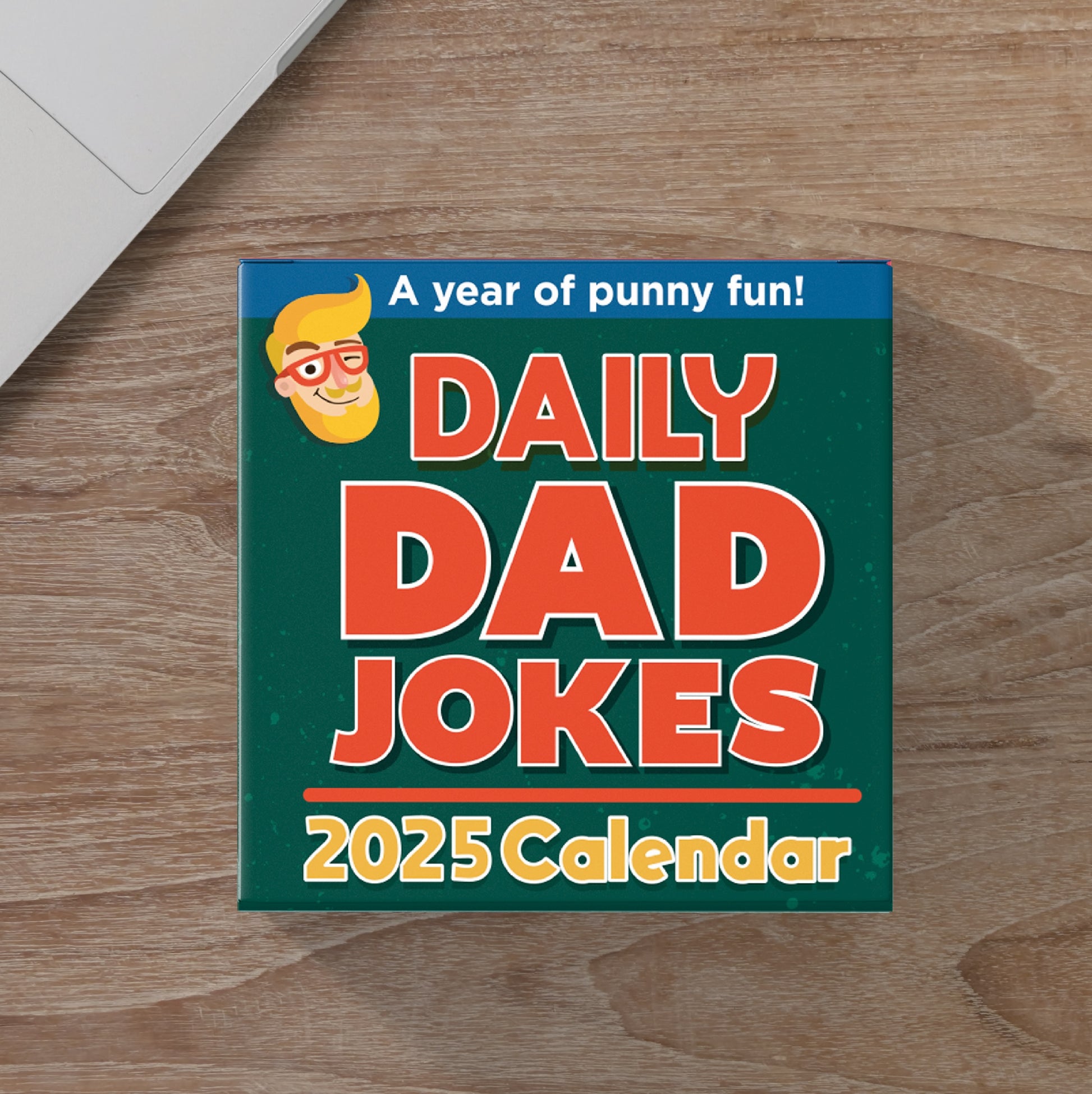 A box of the "2025 Dad Joke Gift Set" by That Is So Dad, featuring a desktop calendar tagged with "A year of punny fun!" and complemented by a dad joke button, sits on a wooden surface next to the corner of a laptop.