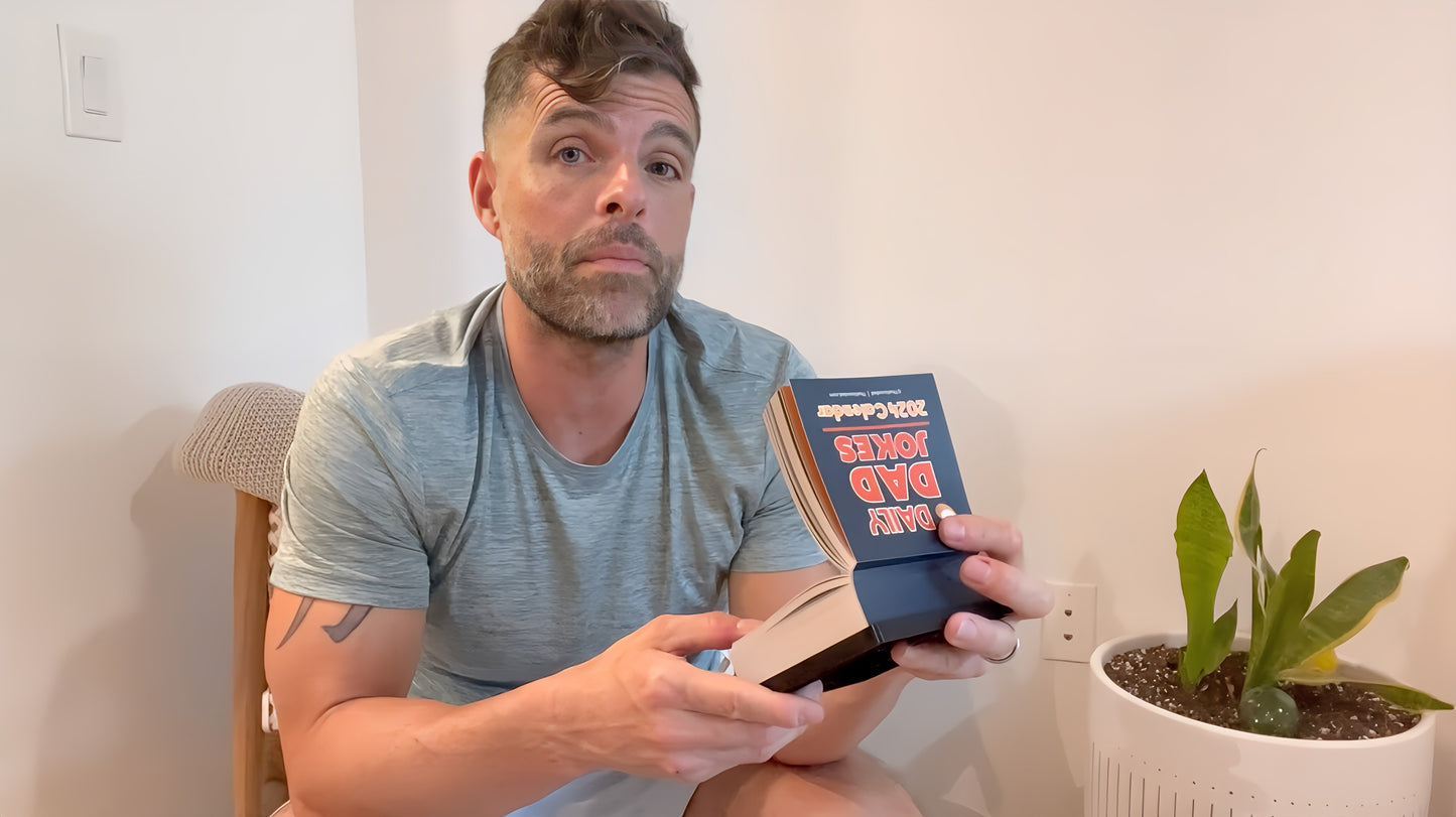 A man wearing a gray shirt holds a "2025 Dad Joke Desktop Calendar" by That Is So Dad while seated next to a potted plant, flipping through pages filled with funny dad jokes perfect for any major world holidays.